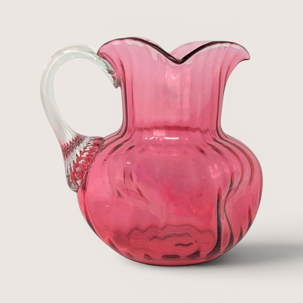 A pink cranberry art glass pitcher with a ribbed body and a clear applied glass handle on a beige backdrop