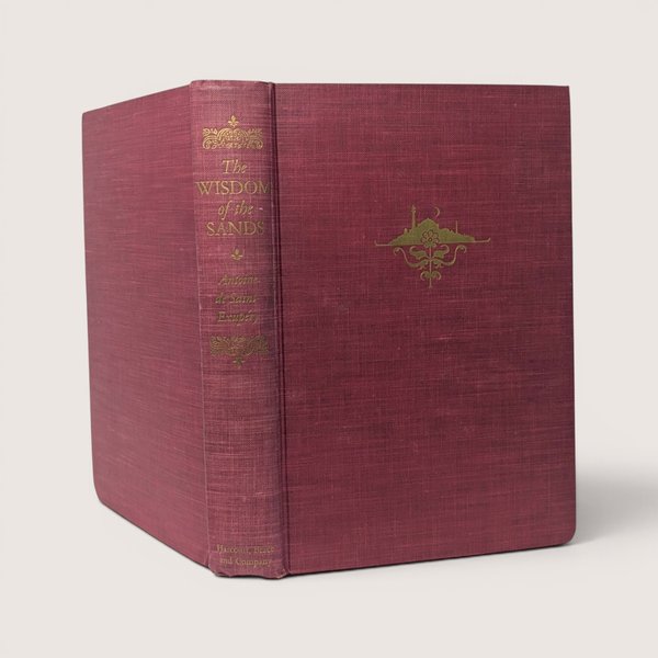 1950 vintage red cloth hardcover copy of The Wisdom of the Sands by Antoine de Saint-Exupéry standing upright against a plain linen background.