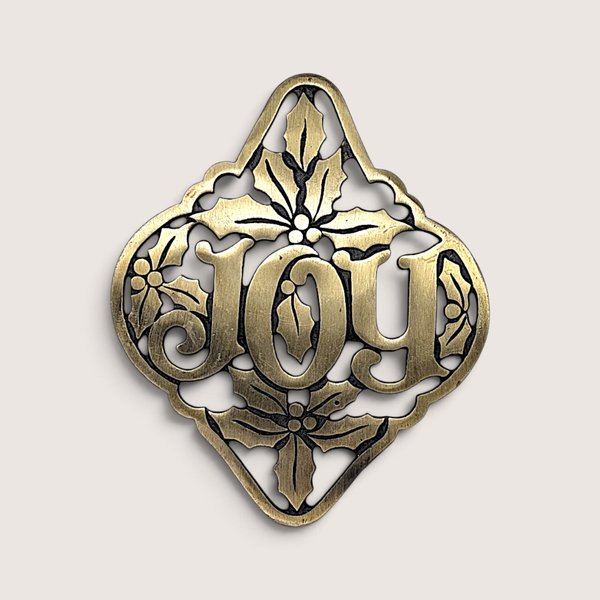 Vintage cast brass holiday trivet featuring openwork "JOY" typography framed by sculpted holly leaves and berries.