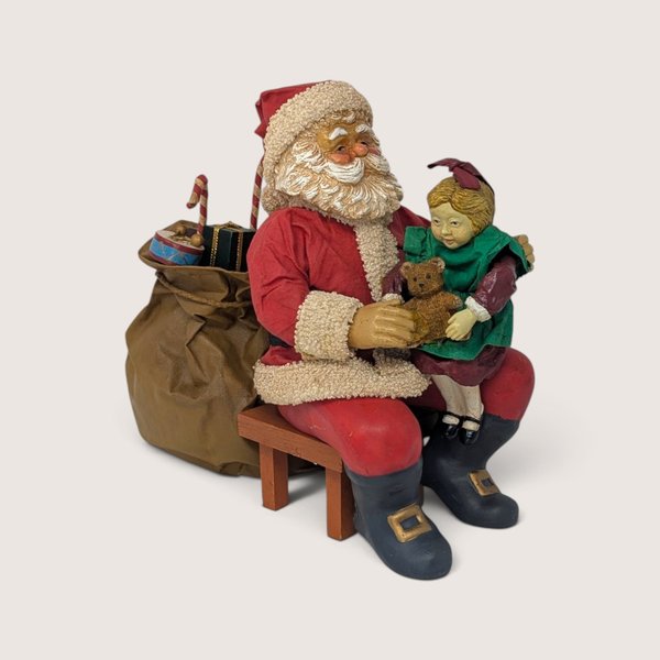 Vintage holiday figurine of Santa Claus sitting on a wooden bench holding a young girl on his lap.
