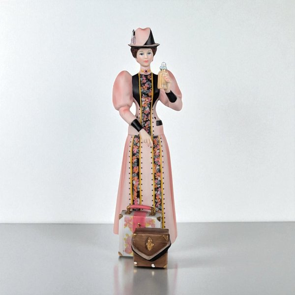 Hand-painted porcelain figurine of a woman in a cream Victorian traveling suit with blue trim and a matching hat. She stands next to a large brown suitcase and travel bag, holding a round hat box and a small purse. The finish is a soft, matte bisque.