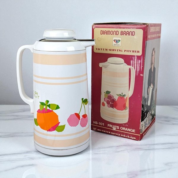 A tall, cream-colored Diamond Brand vacuum pitcher features a vibrant graphic of an orange and pink cherries. It has a white handle, a chrome-finished spout, and sits beside its original red and white box showing a 1970s-style floral fruit motif.