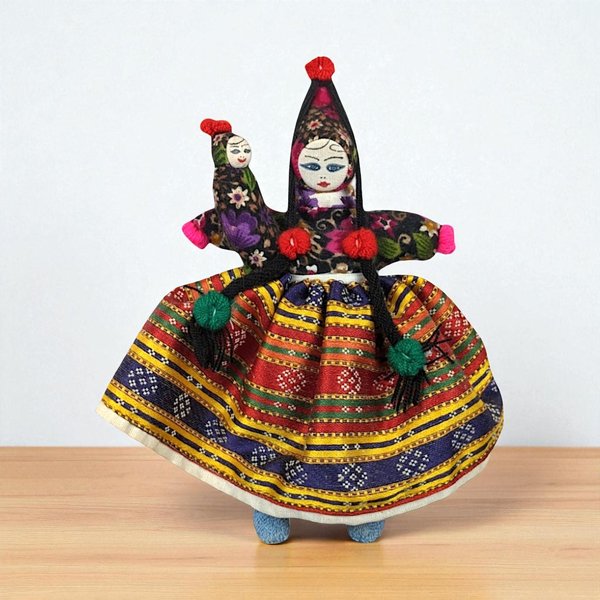 A 9.25-inch tall handmade folk art doll featuring a woman with long black braids holding a smaller baby doll, wearing a multi-colored striped skirt and a tall floral headpiece.