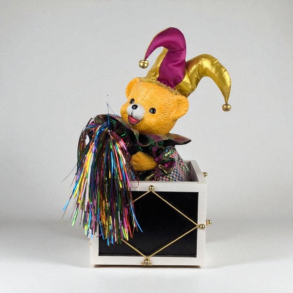 A textured yellow bear cub wearing a purple and gold jester hat with small bells. The bear emerges from a black and white harlequin box, holding a bundle of multi-colored iridescent tinsel. The piece features a mix of smooth plastic and shimmering fabric.