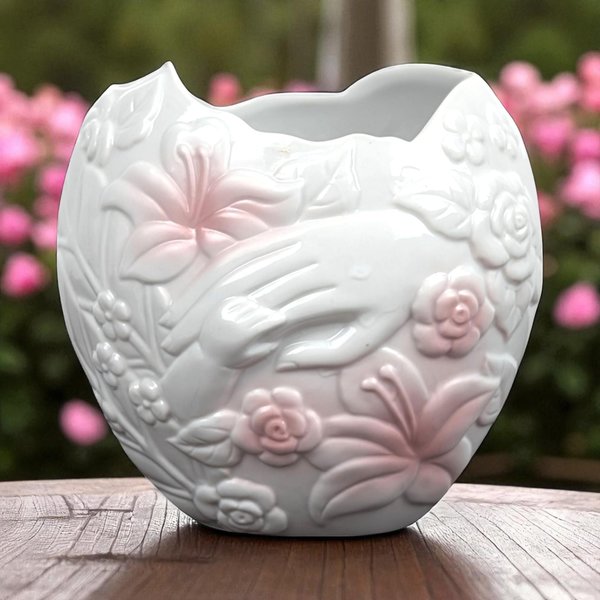 A 5.5-inch tall white porcelain vase with a scalloped rim, featuring embossed 3D designs of a mother’s hand and a small child’s hand surrounded by pink-tipped lilies and roses.