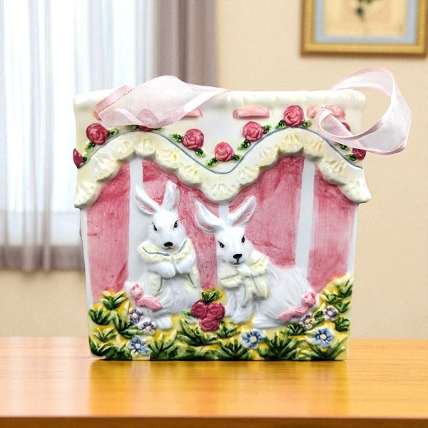 A square ceramic vase shaped like a gift bag features a dimensional relief of two white rabbits in a garden. It is decorated with pink stripes, a scalloped yellow lace border, and tiny red roses, finished with a sheer pink ribbon handle.