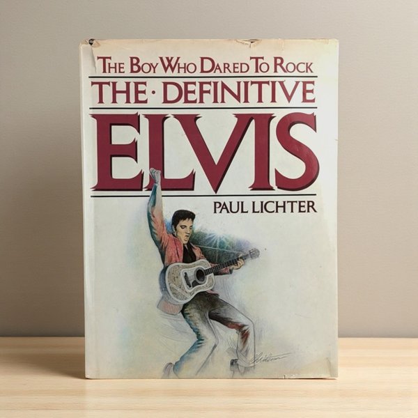 A vintage hardcover book with a cream-colored dust jacket featuring a stylized illustration of Elvis Presley in a pink jacket, mid-performance with a guitar. Large maroon serif text dominates the top half of the cover against a clean, minimalist background.