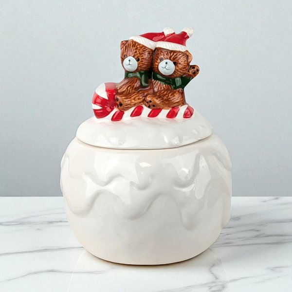 A glossy ceramic jar shaped like a snow-covered mound. The lid features two brown bears wearing red Santa hats and green scarves, sitting atop a red and white striped candy cane. The base has a dripping white glaze texture.