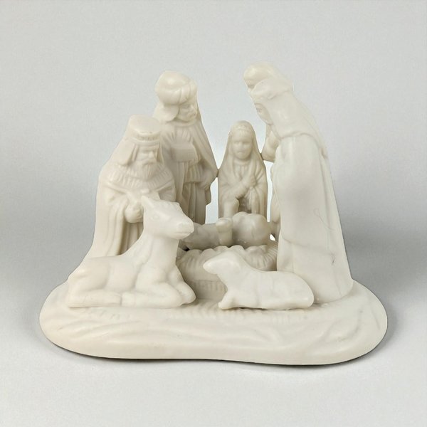A monochromatic white porcelain nativity scene featuring a smooth, matte texture. The sculpture depicts a group of figures and animals on a rounded base, including the Holy Family and Wise Men, characterized by soft silhouettes and refined, uniform coloring.