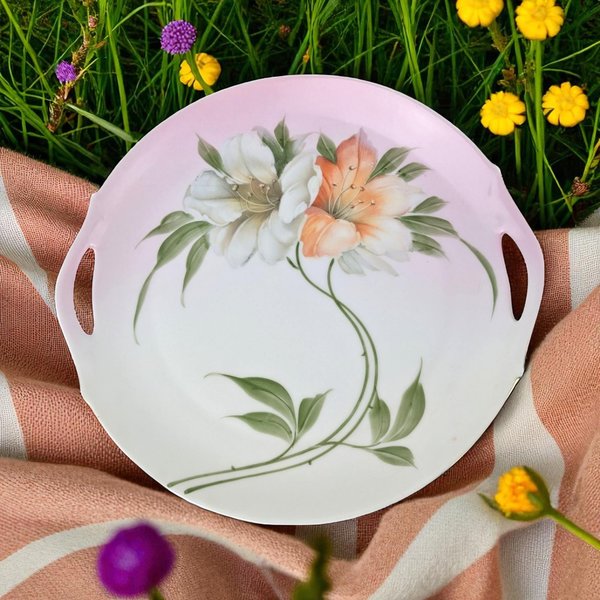 A circular porcelain serving tray featuring two small, integrated side handles. The center is decorated with a hand-painted floral design of one white and one orange lily with green leaves, set against a soft pink and white gradient background.