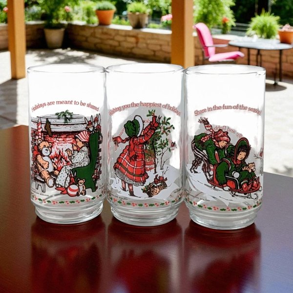 A set of three clear glass tumblers featuring vintage Holly Hobbie illustrations in red, green, and brown. The designs depict nostalgic holiday scenes: a child by a fireplace with a teddy bear, a figure decorating a tree, and two children on a sled, each accompanied by a festive sentiment in red script.