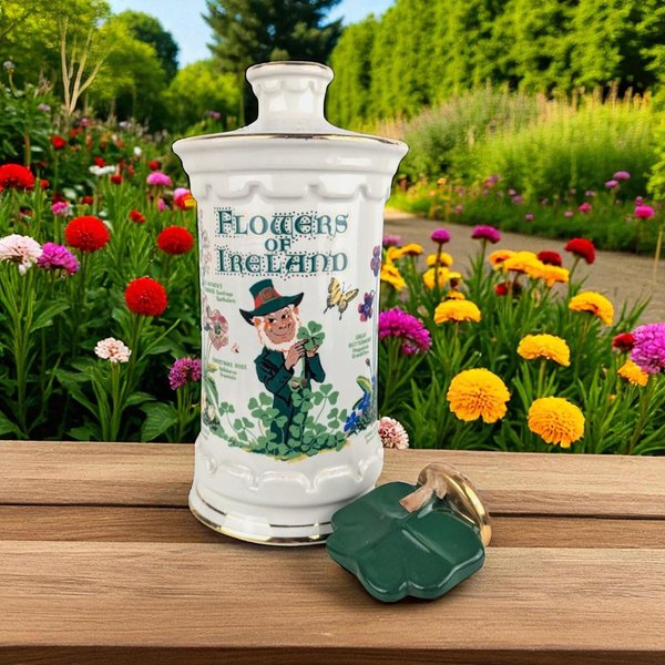 White ceramic apothecary-style canister with gilded trim. Features a "Flowers of Ireland" botanical design with a central leprechaun illustration and floral sprigs. A dark green shamrock-shaped lid sits on a wooden surface beside the jar.