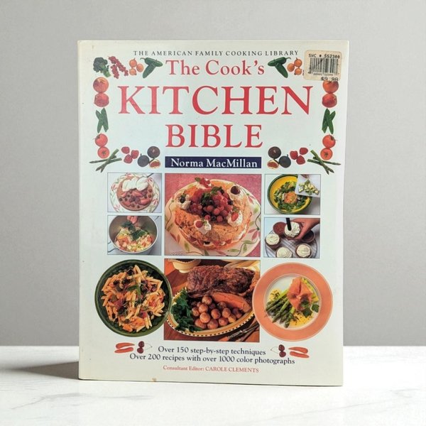 A large hardcover cookbook titled "The Cook's Kitchen Bible" by Norma MacMillan. The front cover features a grid of colorful food photography including a berry-topped cake, pasta, and roasted meat, framed by a border of illustrated fruits and vegetables.