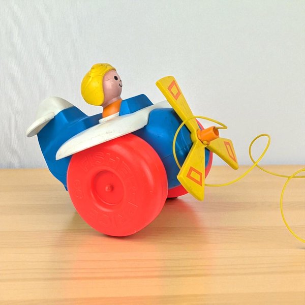 A vintage blue plastic Fisher Price pull toy airplane featuring large red wheels, a yellow three-blade propeller, and a built-in pilot figure with blonde hair and a smiling face.