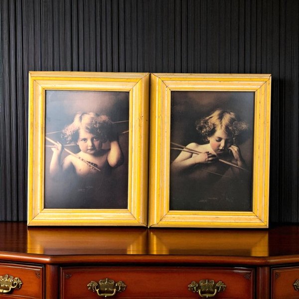 A pair of vintage framed prints featuring a young child posed as Cupid with a bow and arrow, titled "Cupid Awake" and "Cupid Asleep," set in yellow wooden frames.