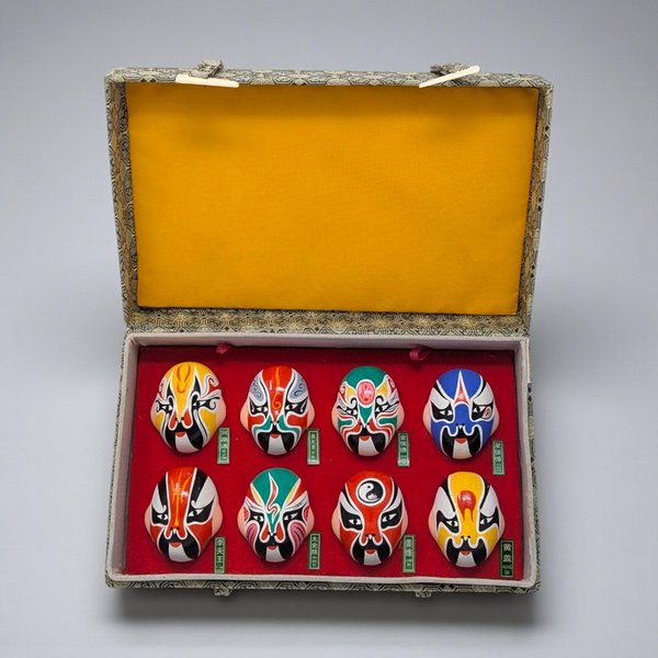Set of eight miniature hand-painted Chinese opera masks displayed in a red velvet-lined box. Each mask features vibrant, high-contrast colors including red, blue, green, and yellow with bold black-and-white patterns and smooth, glossy textures.