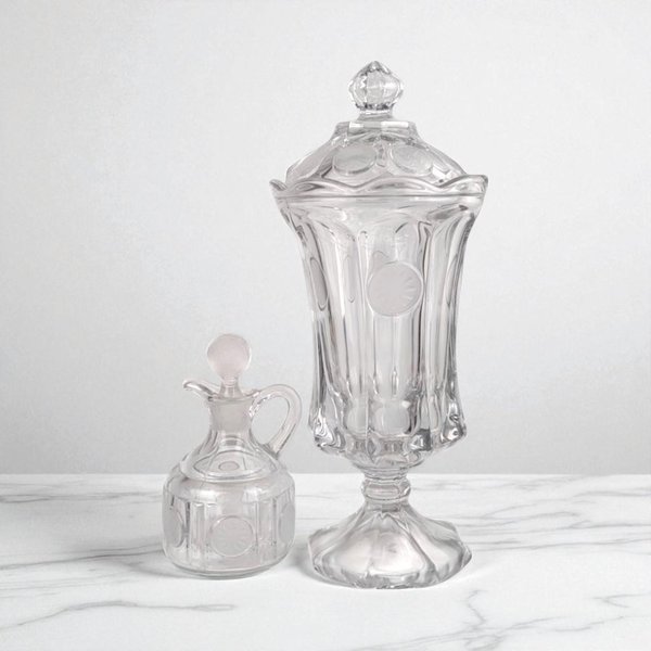 A clear glass apothecary jar with a matching lid stands next to a small glass cruet with a stopper. Both pieces feature a thumbprint pattern and vertical ribbing. They have a sleek, transparent silhouette and polished finish, set against a light marble surface.