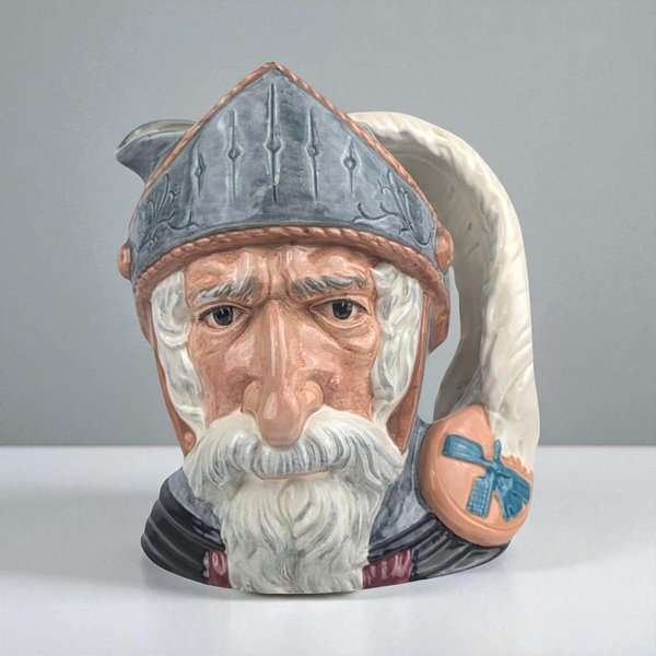 A ceramic character jug depicting Don Quixote. The figure features a weathered face with a white mustache and beard, wearing a grey pointed helmet. A large white plume forms the handle, sweeping down to a small shield featuring a blue windmill.