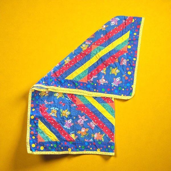 A square, handmade textile throw featuring a repeating pattern of whimsical clown faces. The blanket showcases hand-stitched or appliquéd details with vibrant primary colors like red, yellow, and blue against a neutral background. The edges are finished with a simple hand-bound border.