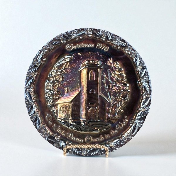 A round carnival glass collector plate with a dark, iridescent amethyst or black finish that reflects metallic purple and bronze tones. The center features a high-relief embossed scene of a church with a tall steeple surrounded by bare winter trees. A decorative holly leaf border encircles the rim.