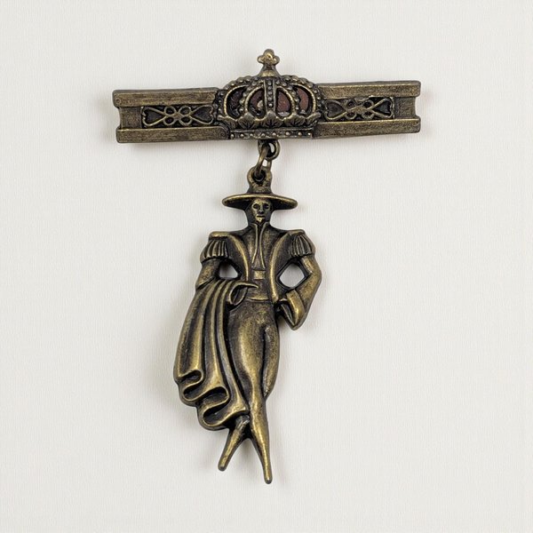 A vintage-style bronze-toned brooch features a detailed figure of a Spanish matador in traditional dress and cape, dangling below a decorative bar pin topped by a matching crown, on a light background.