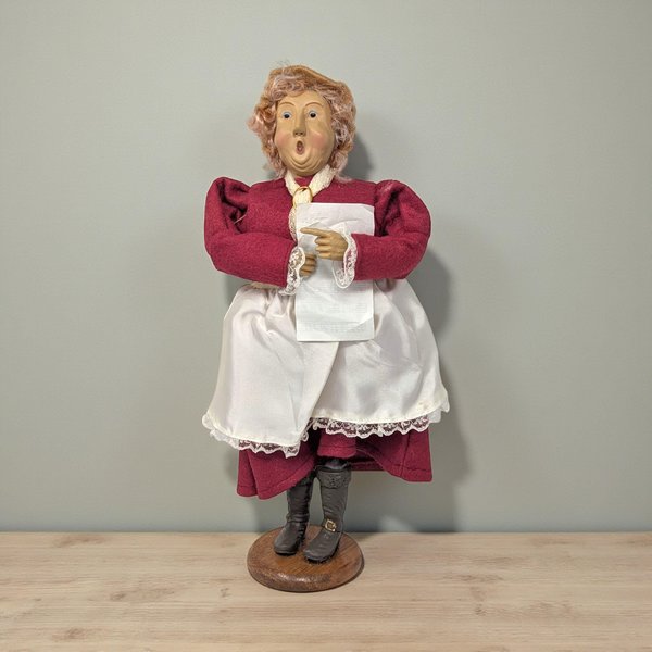 A vintage handcrafted caroler figurine of a woman with a surprised, singing expression and curly reddish-brown hair. She wears a deep burgundy dress under a white satin apron with lace trim, holding a sheet of music and standing on a round wooden base.