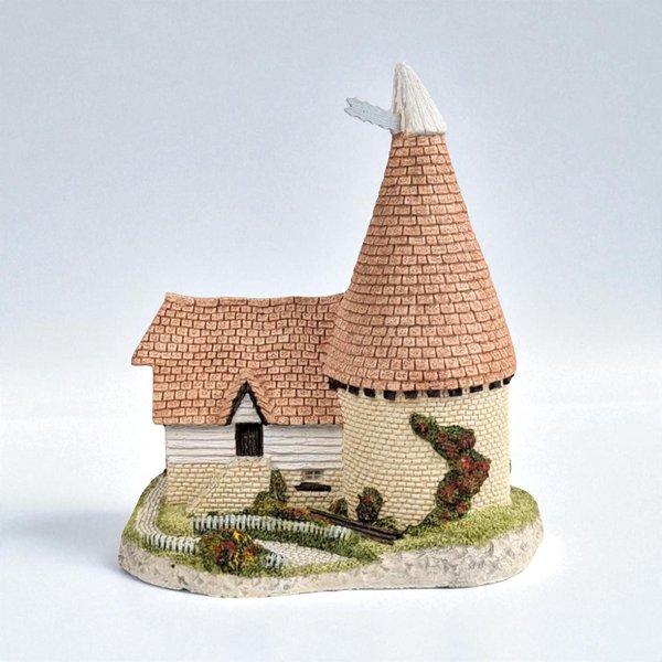 A hand-painted miniature cottage figurine with a tall, conical tiled roof. The structure features detailed cream brickwork, a small white wooden annex, and climbing red flowers along the circular tower, all set on a textured stone base with a tiny picket fence.