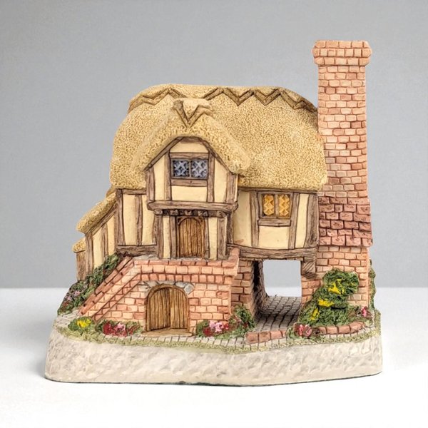 A hand-painted miniature thatched-roof cottage with a large, square brick chimney and half-timbered walls. The figurine includes a textured stone base with a tiered brick entryway, a small arched wooden door, and a pass-through opening under the main floor.