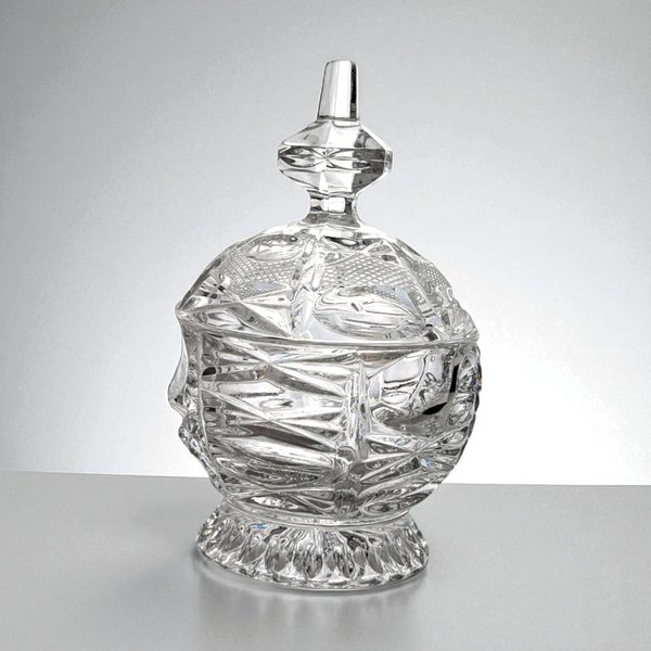 A heavy, clear cut-glass candy dish with a matching lid. The vessel features deep, geometric diamond and starburst patterns that create a prismatic effect. The lid is topped with a tiered, pointed finial, and the entire piece sits on a short, flared pedestal base with a scalloped edge.