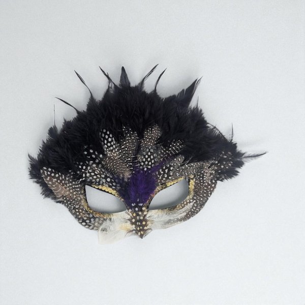 A masquerade eye mask featuring a layered arrangement of real feathers. A crown of soft black plumes sits atop speckled grey and white guinea fowl feathers. Purple down accents the bridge, while gold sequins trim the eye cutouts.