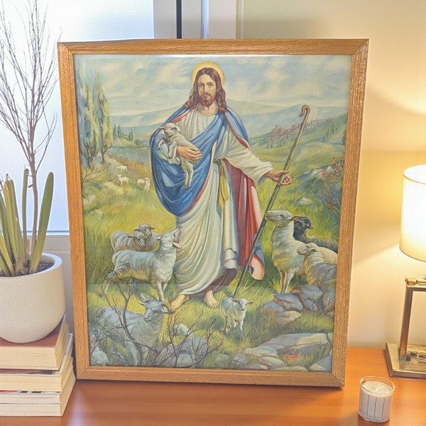 A framed religious lithograph featuring Jesus as the Good Shepherd. He stands in a rolling green landscape wearing white and blue robes, cradling a small lamb while surrounded by a flock. The piece is housed in a simple, light-toned wood frame with a visible grain.