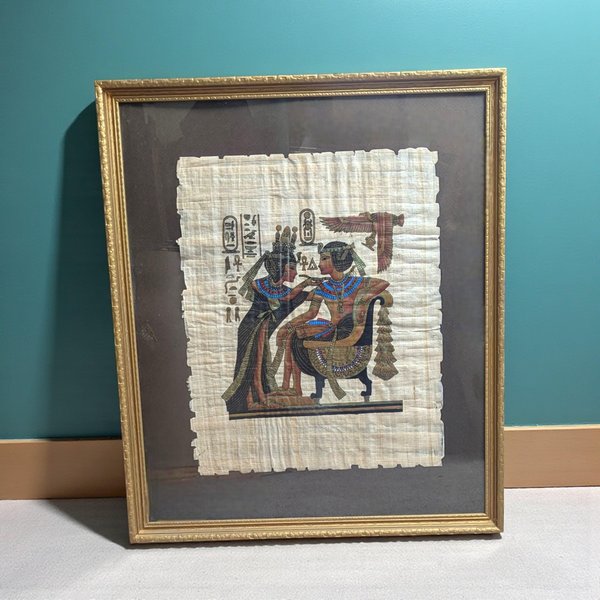 A rectangular Egyptian papyrus painting with deckled edges mounted on a dark brown mat. The scene depicts two figures in traditional ancient Egyptian attire and jewelry, accompanied by hieroglyphics and a winged deity. The piece is housed in a gold-finished frame.