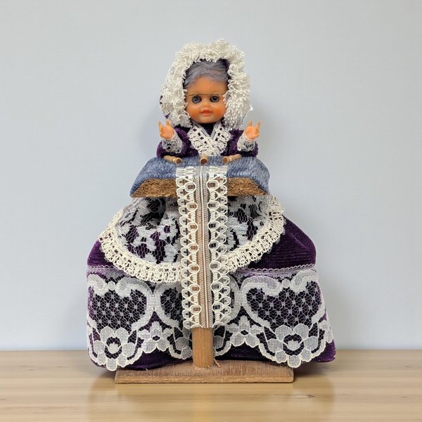 A small vintage doll depicting a lacemaker. She wears a deep purple velvet dress and a white ruffled bonnet. A white lace apron and trim overlay the skirt. She sits behind a wooden stand with a blue velvet pillow and lace-making bobbins.