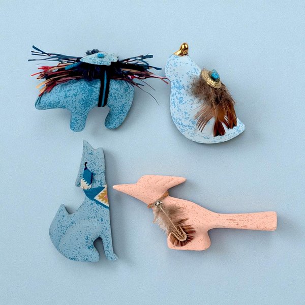 A horizontal view of four small, speckled ceramic animal figurines arranged on a white surface. The set includes three blue-speckled figures—a bear, a bird with brown feathers and a turquoise accent, and a sitting wolf wearing a patterned bandana. A pink-speckled roadrunner with a small brown feather and a tiny bell collar stands in the foreground.