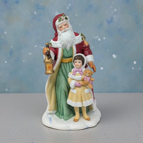 Matte porcelain figurine of Santa in a red and green robe holding a gold lantern. A young girl in a yellow winter coat stands beside him holding a teddy bear on a snowy base.