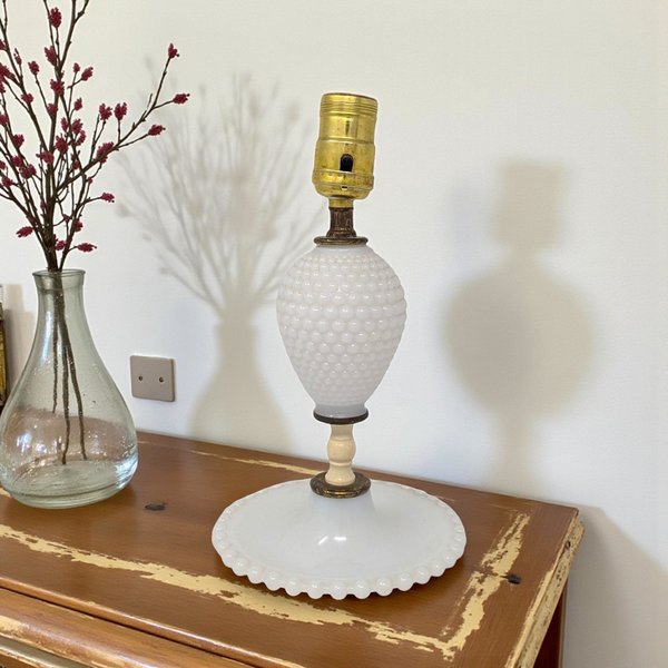 White milk glass vanity lamp featuring a hobnail texture on the central ovoid body and a ruffled, beaded edge along the wide circular base. The lamp has aged brass hardware and a cream-colored spindle neck, creating a slender, upright silhouette.