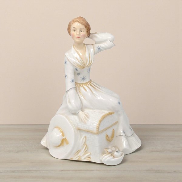 Glossy porcelain figurine of a woman in a white gown accented with blue starbursts and gold trim. She sits atop a textured trunk with a wide-brimmed hat at her side. The piece features a smooth, reflective finish and a graceful, seated silhouette.