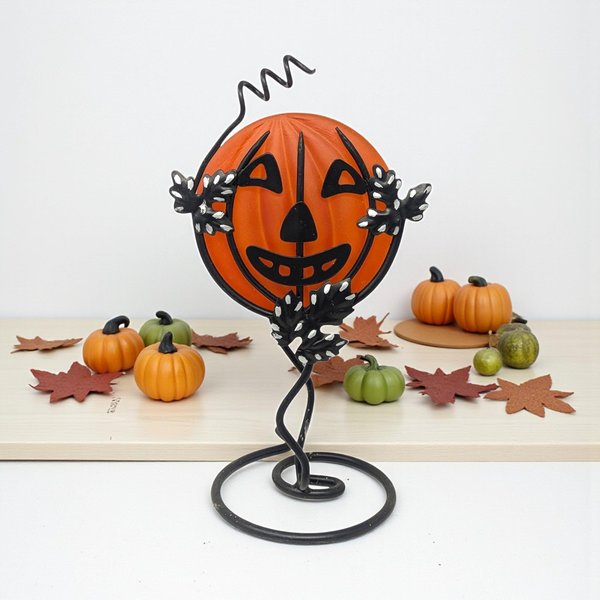 A frosted orange glass pumpkin face is cradled within a black wrought iron stand. The metal frame features a coiled base, climbing vine-like tendrils, and speckled leaves. A whimsical wire curl extends from the top of the jack-o'-lantern silhouette.