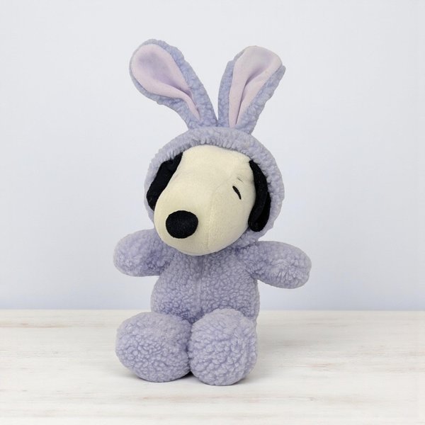 Small plush Snoopy dog wearing a textured, lavender-purple bunny costume. The hood features tall rabbit ears with pale pink interiors. Snoopy has a white face, black nose, and closed, peaceful eyes. The body is a soft, sherpa-style fleece.