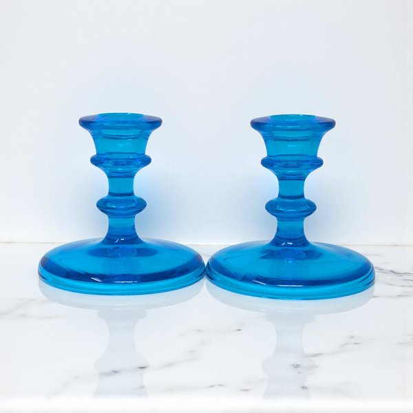 A pair of vibrant cerulean blue glass taper candle holders featuring a tiered, sculptural stem and a wide, circular disc base. The translucent glass has a smooth, glossy finish and a symmetrical silhouette.
