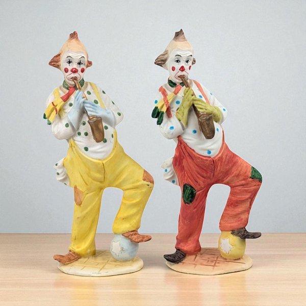 Two ceramic clown figurines standing in mirrored poses, playing saxophones. One wears yellow trousers and the other red, both with polka-dot shirts and patches. Their silhouettes feature one leg raised on a star-patterned ball with a matte, hand-painted texture.