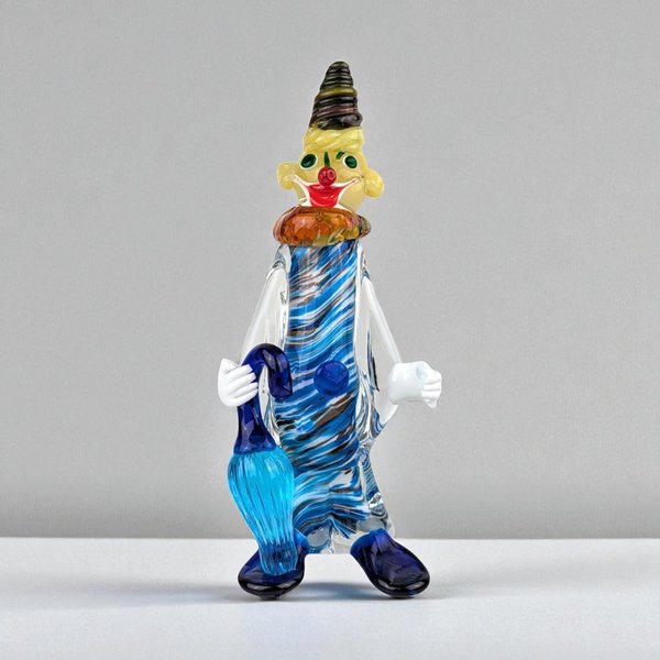 A hand-blown glass clown figurine features a slender, translucent body with internal blue and white swirls. He holds a bright blue umbrella and wears a pointed brown hat, an orange ruffled collar, and oversized navy blue shoes.