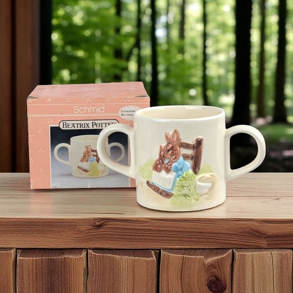 A cream ceramic two-handled child's mug featuring a raised, 3D relief of a rabbit in a blue coat. The smooth, glossy vessel sits beside its original pink Schmid box on a wooden surface.