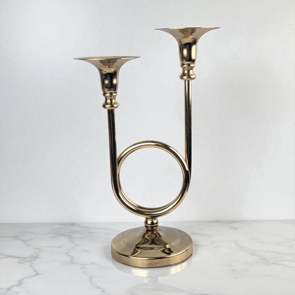 Polished brass double candle holder featuring a central circular ring motif. Two slender vertical arms of differing heights rise from the ring to hold tulip-shaped cups. The finish is a bright, reflective gold with a smooth, metallic texture and a wide pedestal base.