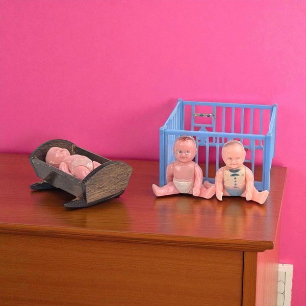 Three small, pink plastic baby dolls. One rests in a dark wood-grain rocking cradle. Two sit in front of a light blue slatted plastic playpen. Smooth, molded textures with painted blue and white diaper details.