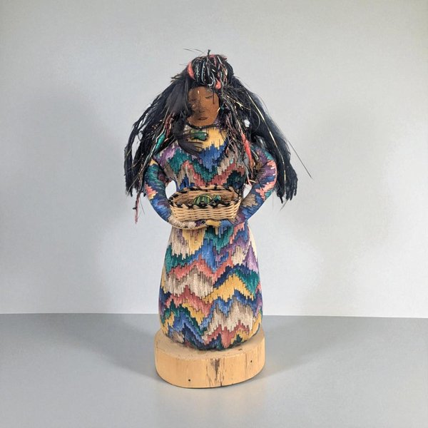 Handcrafted folk-art doll with long black fiber hair and a vibrant chevron-patterned dress in blue, pink, and gold. She holds a small woven basket and stands on a natural wood base. The texture is a mix of coarse textile and smooth wood.
