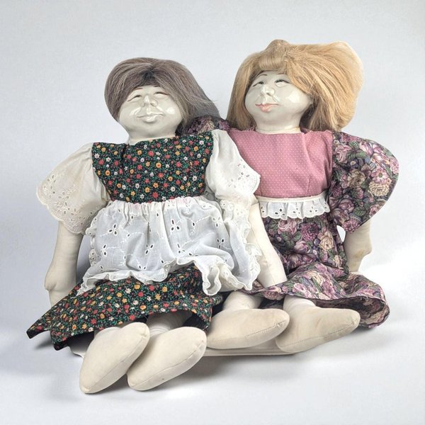 Pair of handcrafted textile dolls with detailed ceramic faces, sitting side-by-side. One has grey hair and a floral print dress with a white eyelet apron. The other has blonde hair and a pink bodice with a purple floral skirt. They are on a simple grey background.