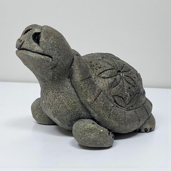 A small, heavy-textured stone turtle figurine with a weathered grey finish. The shell is embellished with a carved floral or sand dollar motif, and the figure is depicted with an upturned head and simple, rounded limbs.