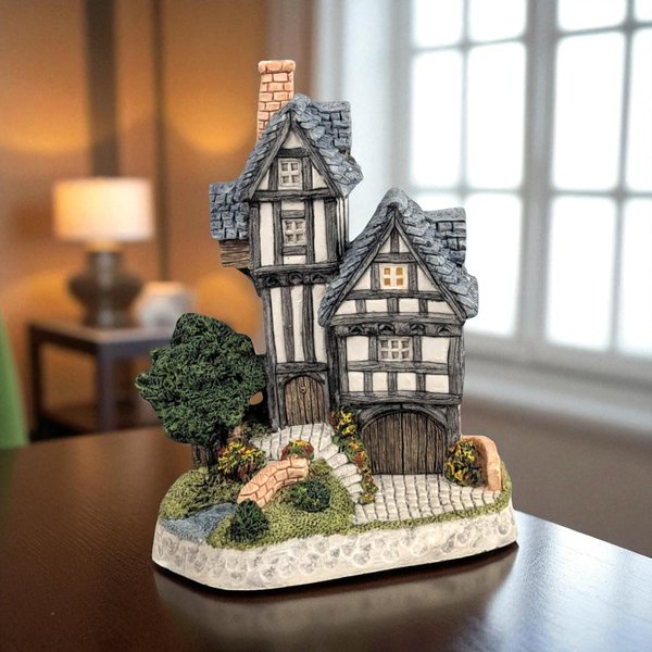 A hand-painted miniature Tudor-style cottage featuring a prominent, vertical timber-framed tower and a smaller gabled wing with slate-grey roof tiles. The figurine includes a textured stone base with a winding path, small shrubs, and a miniature stone bridge over a blue stream.