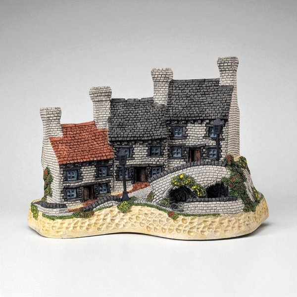 A hand-painted miniature stone cottage row with three distinct sections featuring white brickwork, grey and red shingled roofs, and four prominent chimneys. The figurine includes a textured stone base with a winding path, a stone bridge, and two black streetlamps.